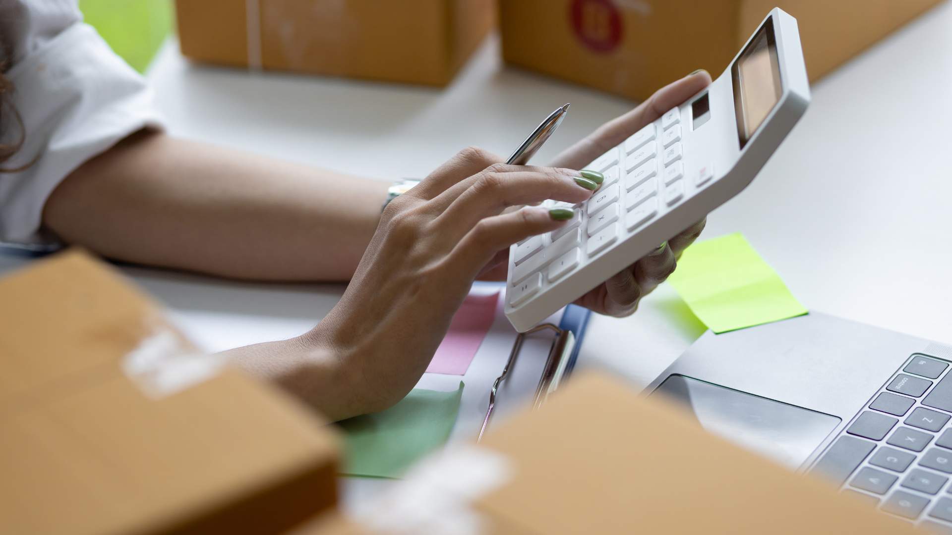 A hand holding a calculator and pen with a laptop and green sticky note in front of them on a desk.