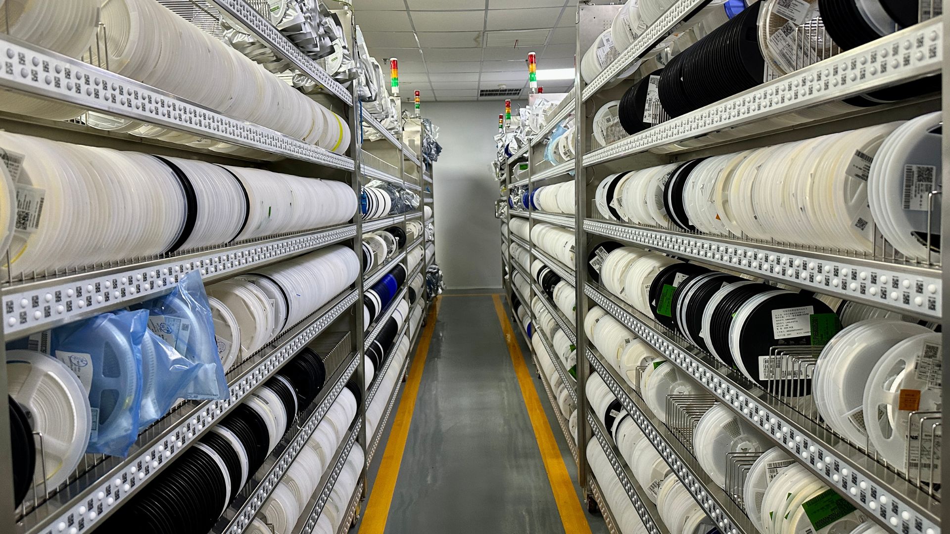 A narrow warehouse aisle lined with shelves on both sides, filled with reels of components and electronic parts, with labeled spools organized in rows.