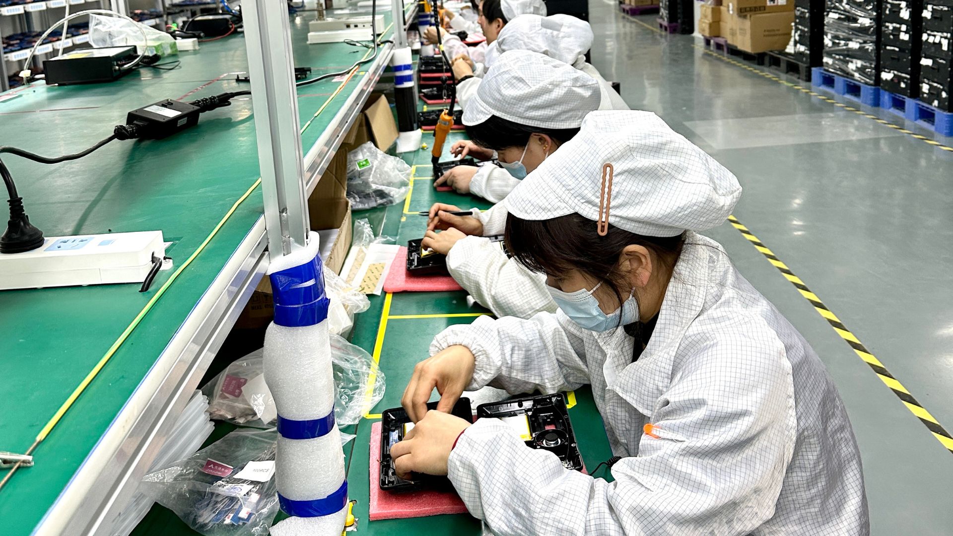 Workers in protective clothing and face masks assembling electronic devices at a production line in a manufacturing facility.