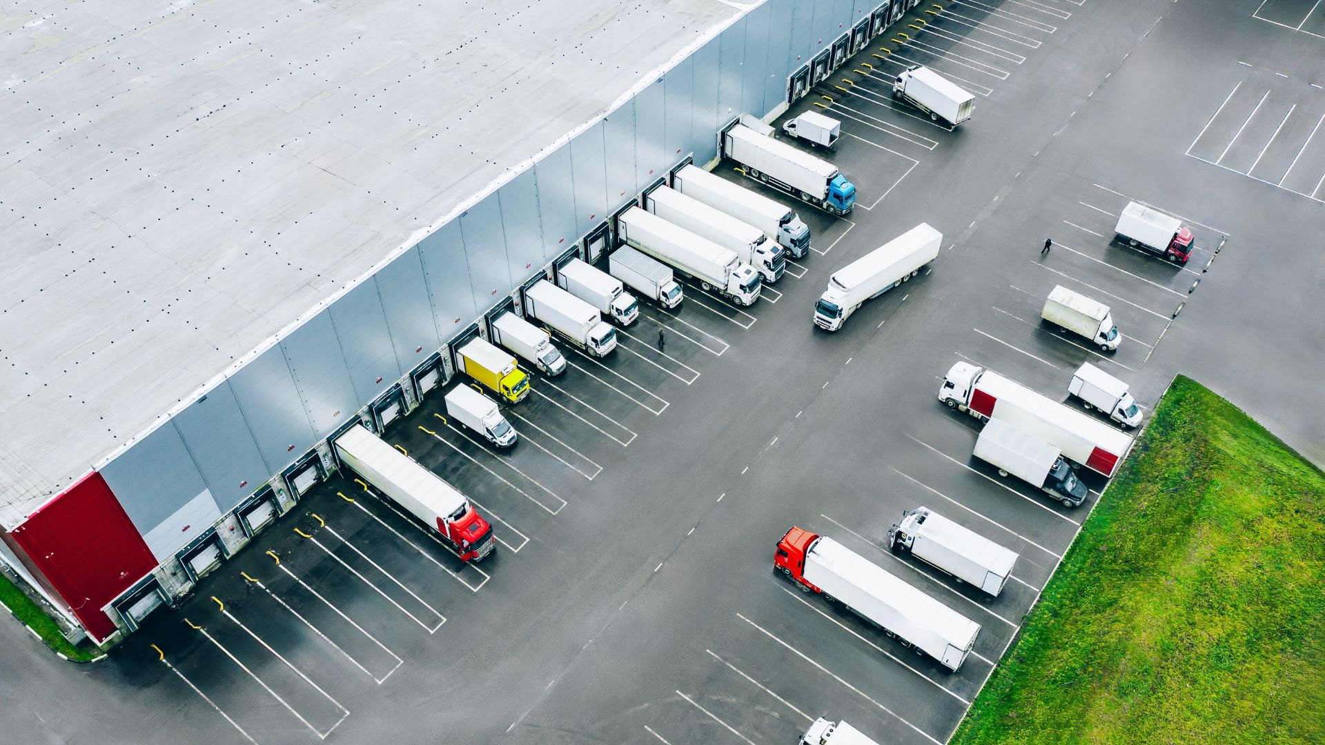 A warehouse parking lot with the edge of the warehouse in view, featuring several vans and lorries.