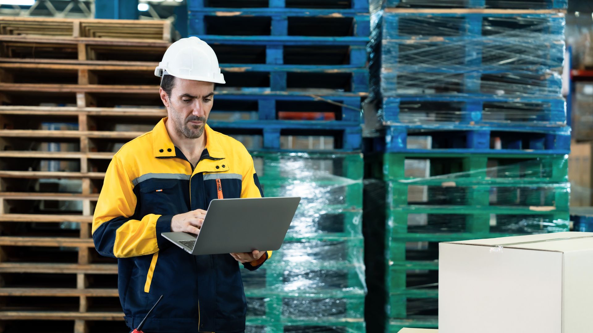 A pallet pooler on a laptop standing in front of large stacks of wrapped reusable plastic pallets.