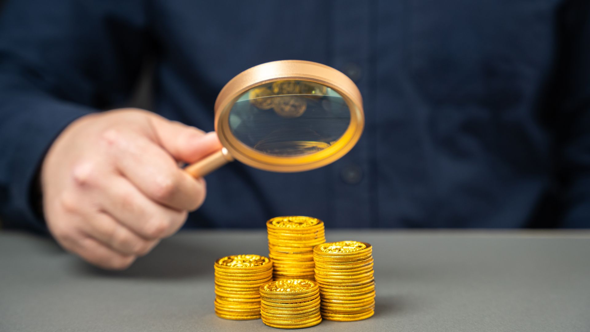 A close-up of someone holding a magnifying glass over stacks of gold coins.