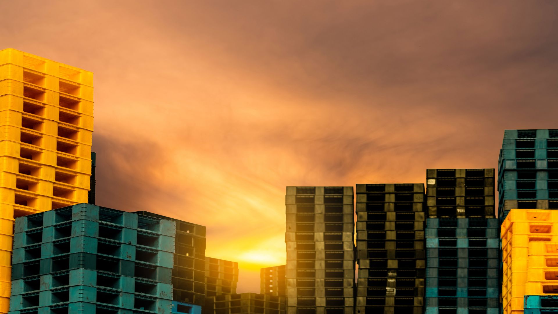 Teal and yellow plastic pallets stacked up tall outside in front of a bright sunset.