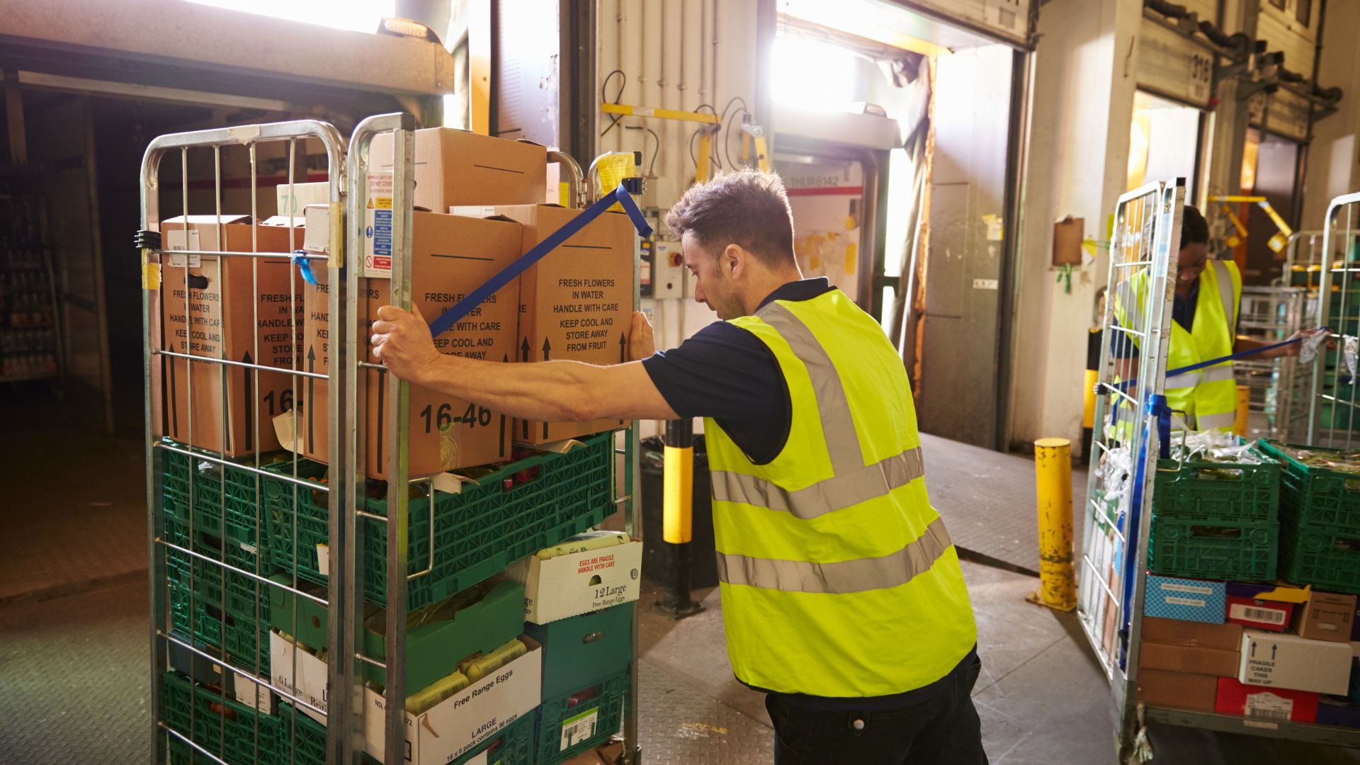 A person wearing a high-vis jacket pushing a roll cage in a warehouse.