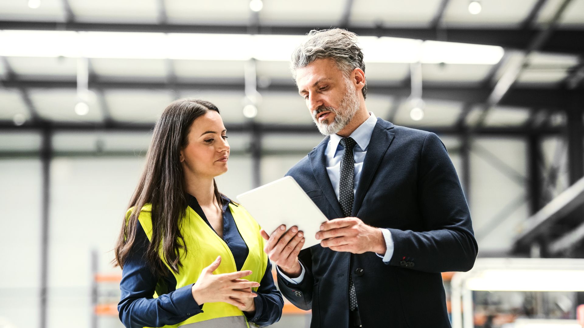 Two logistics professionals, one in a hi-vis jacket and one in a suit, talking and holding a tablet.